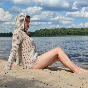 May include: A light beige, open-weave crochet hooded sweater with long sleeves. The sweater is worn on a sandy beach with a lake and trees in the background. The sweater has a circular design on the front.