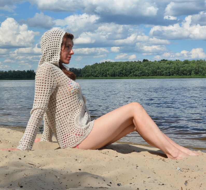 May include: A light beige, open-weave crochet hooded sweater with long sleeves. The sweater is worn on a sandy beach with a lake and trees in the background. The sweater has a circular design on the front.