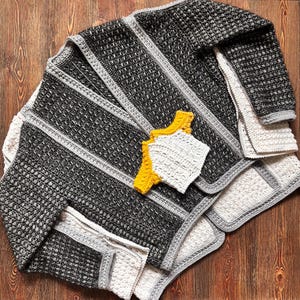 May include: A handmade crochet blanket in shades of black, gray, and white, with a textured stitch pattern and gray border. Accompanying the blanket are two matching coasters, one white and one yellow. The blanket is folded on a wooden surface.