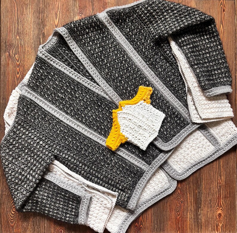 May include: A handmade crochet blanket in shades of black, gray, and white, with a textured stitch pattern and gray border. Accompanying the blanket are two matching coasters, one white and one yellow. The blanket is folded on a wooden surface.