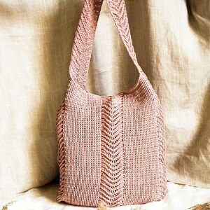 May include: A dusky pink knitted tote bag with a shoulder strap. The bag has a central chevron pattern and is made of a textured, woven material. The bag is shown against a neutral background.