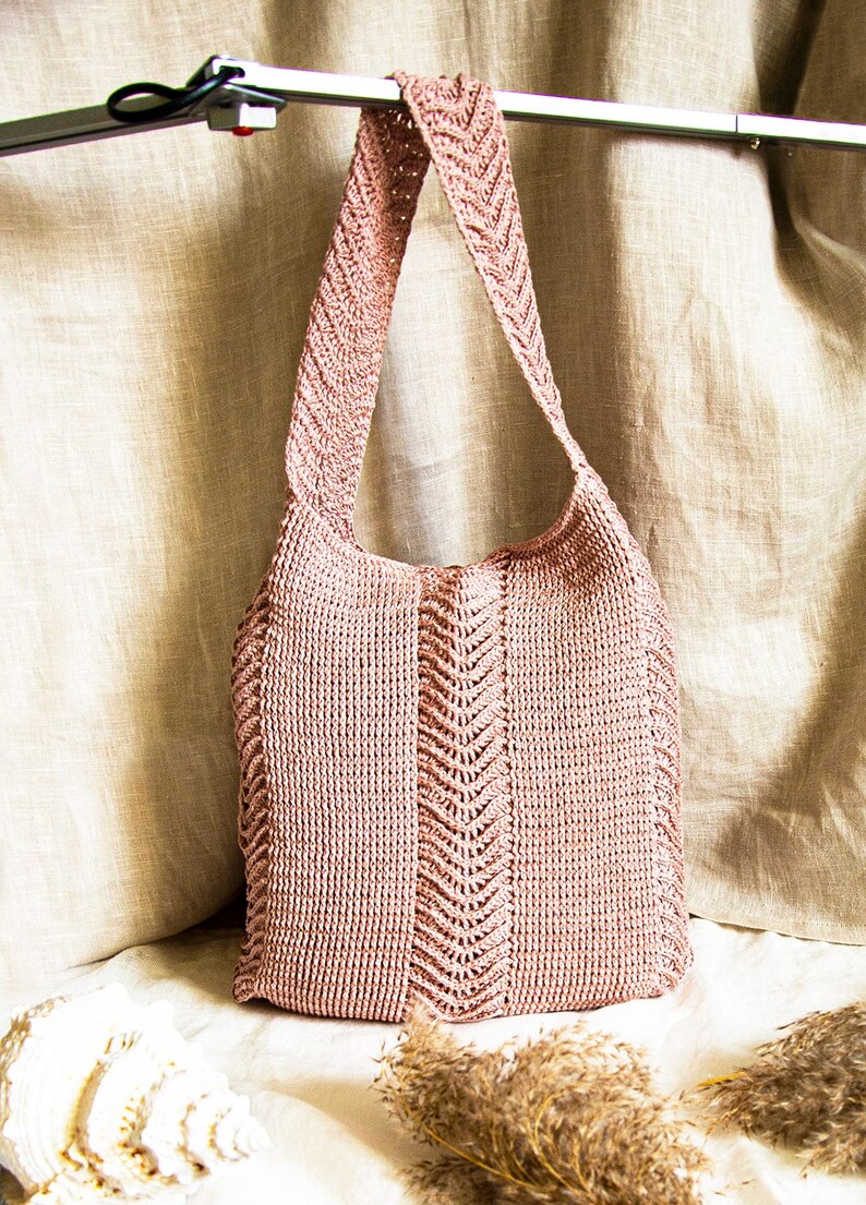 May include: A dusky pink knitted tote bag with a shoulder strap. The bag has a central chevron pattern and is made of a textured, woven material. The bag is shown against a neutral background.
