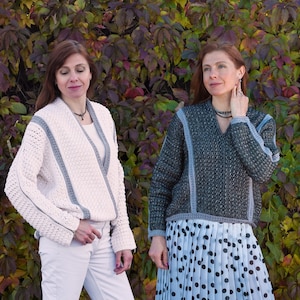 May include: Two women wearing handmade sweaters. One sweater is off-white with gray trim, the other is dark gray with gray trim. The sweaters have a V-neck design and are made of a textured knit material. The background is a colorful array of autumn leaves.