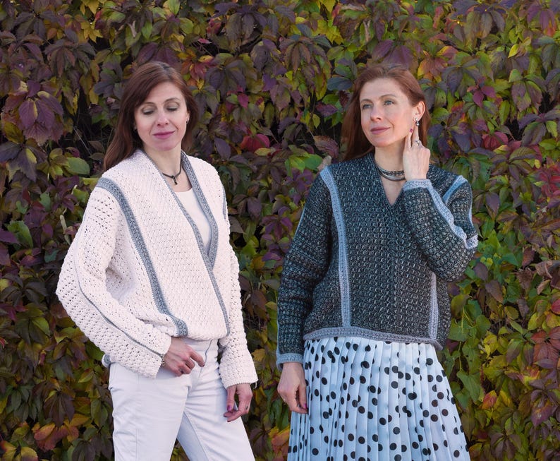 May include: Two women wearing handmade sweaters. One sweater is off-white with gray trim, the other is dark gray with gray trim. The sweaters have a V-neck design and are made of a textured knit material. The background is a colorful array of autumn leaves.