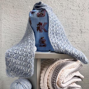 May include: A light blue crocheted scarf draped over a blue pillow with a bear and letter "K" design. A folded blanket with white, gray, and brown stripes sits below, along with a ball of blue and white yarn. The items are on a gray shelf.