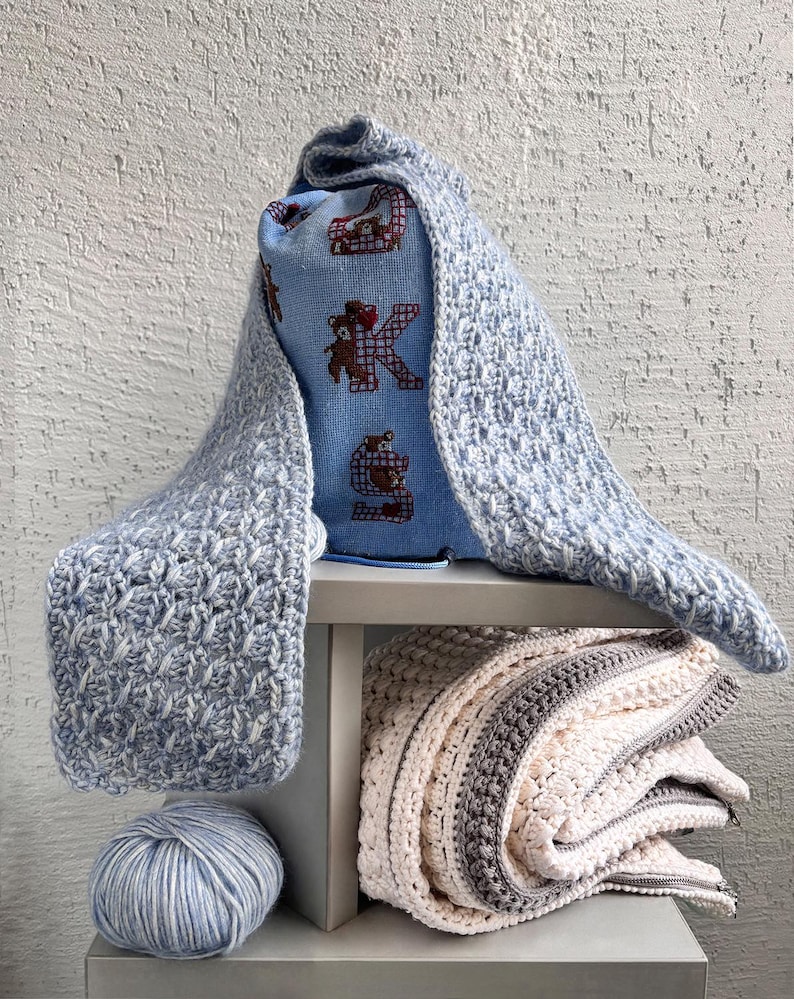 May include: A light blue crocheted scarf draped over a blue pillow with a bear and letter "K" design. A folded blanket with white, gray, and brown stripes sits below, along with a ball of blue and white yarn. The items are on a gray shelf.