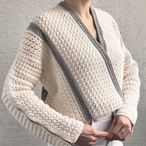 May include: Cream-colored, textured crochet jacket with gray trim. The jacket features a wrap-style design, long sleeves, and a zipper detail on the sleeve. The jacket is a stylish outerwear piece.