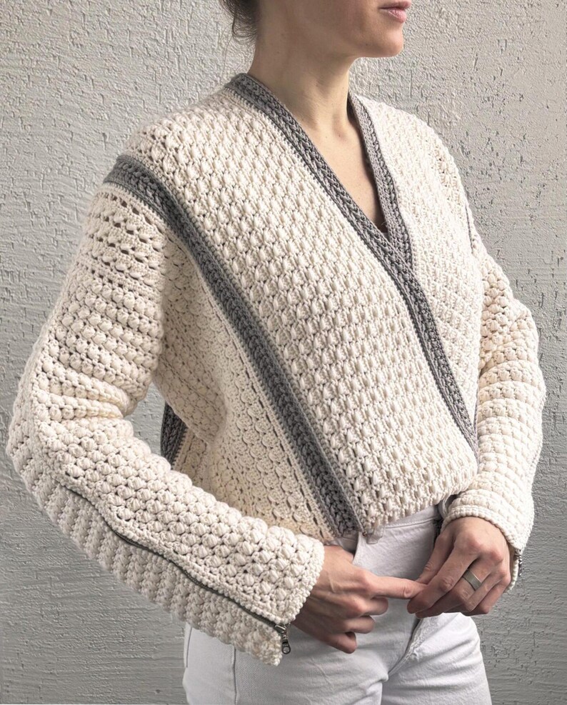 May include: Cream-colored, textured crochet jacket with gray trim. The jacket features a wrap-style design, long sleeves, and a zipper detail on the sleeve. The jacket is a stylish outerwear piece.