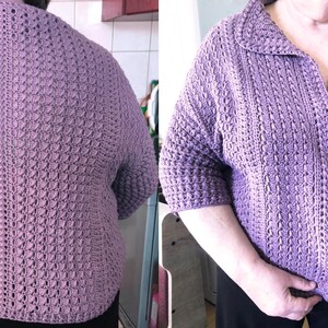 May include: A lavender-colored, crocheted cardigan with a collar and short sleeves. The front of the cardigan has a button closure. The crochet pattern is visible throughout the garment.
