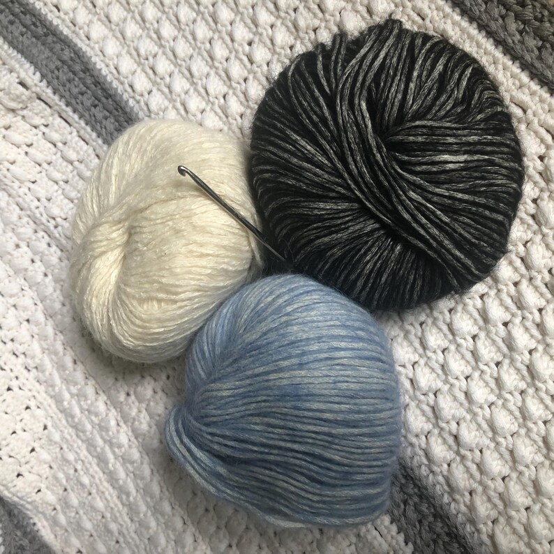 May include: Three balls of yarn in cream, black and gray, and blue. A silver crochet hook rests on the cream yarn. The yarn is on a white and gray textured surface, suggesting a crafting theme.