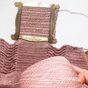 May include: A close-up of pink knitted fabric with a brass gauge tool. The gauge tool is measuring the knitted fabric. A ball of pink yarn is in the foreground. The knitted fabric has a textured appearance.
