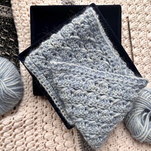May include: A light blue crocheted scarf with a textured pattern, displayed on a black surface. Two balls of light blue yarn and a crochet hook are also visible, suggesting a handmade craft project. The scarf is folded, showcasing its intricate design.