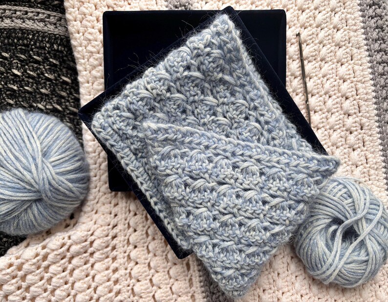 May include: A light blue crocheted scarf with a textured pattern, displayed on a black surface. Two balls of light blue yarn and a crochet hook are also visible, suggesting a handmade craft project. The scarf is folded, showcasing its intricate design.