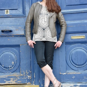 May include: A woman wearing a gray leather jacket, a white crochet top, and black capri pants. She is also wearing beige sandals. The background is a weathered blue door.