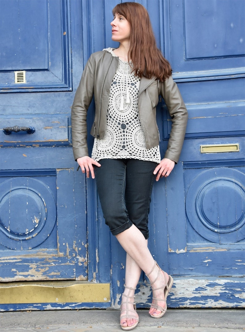 May include: A woman wearing a gray leather jacket, a white crochet top, and black capri pants. She is also wearing beige sandals. The background is a weathered blue door.