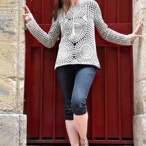 May include: A cream-colored, long-sleeved crochet top with a circular pattern design. The top is paired with dark denim capri pants and strappy sandals. The background features a red door and stone walls.