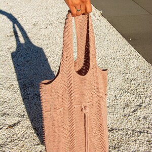 May include: A peach-coloured tote bag with a drawstring closure. The bag is made of a knitted material with a textured pattern. The handles are integrated into the bag's design. The bag is held up by a person.