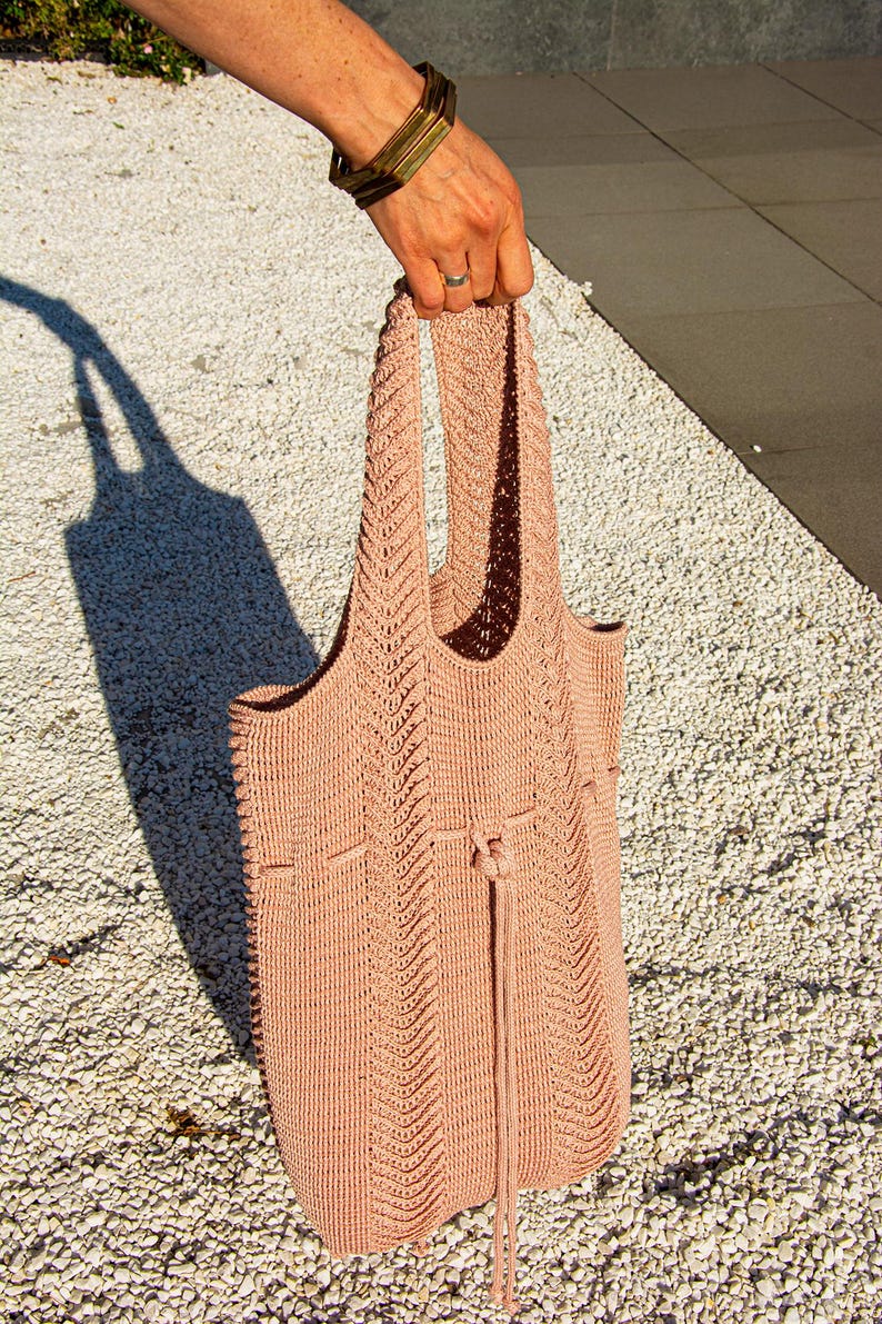 May include: A peach-coloured tote bag with a drawstring closure. The bag is made of a knitted material with a textured pattern. The handles are integrated into the bag's design. The bag is held up by a person.