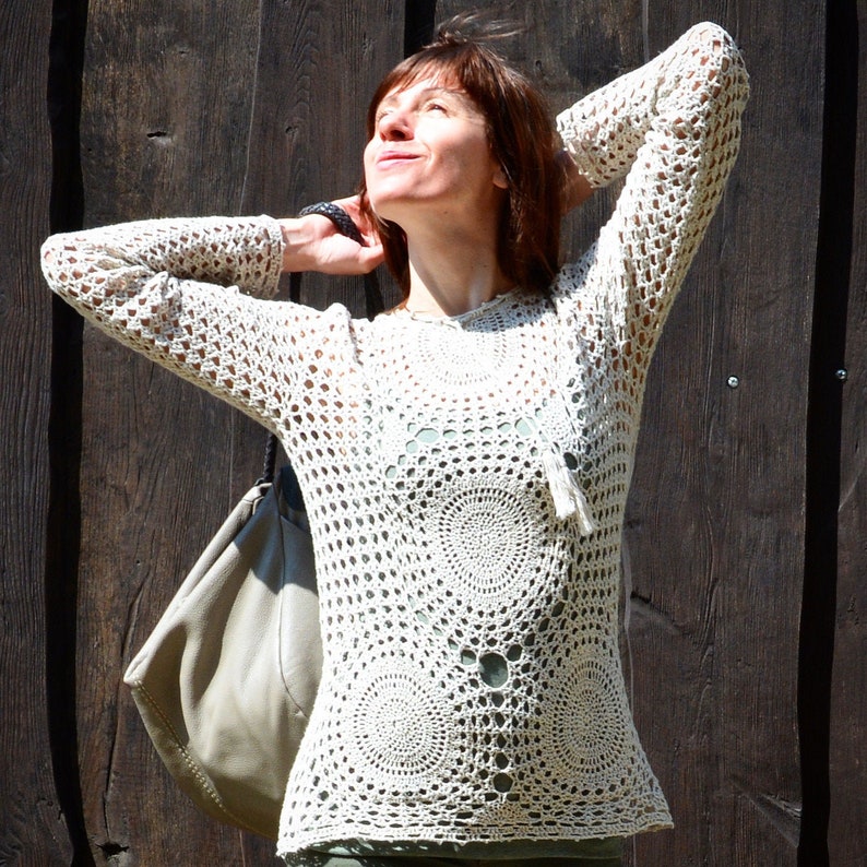 May include: Cream-colored crochet top with long sleeves and circular patterns. The open-weave design reveals a glimpse of the undergarment. The top has a relaxed fit and a tassel detail at the neckline. The background is a dark wooden surface.
