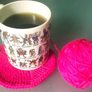 May include: A white mug with a historical figures design, filled with a green liquid, rests on a bright pink crocheted coaster. A ball of bright pink yarn sits nearby, suggesting a crafting theme.
