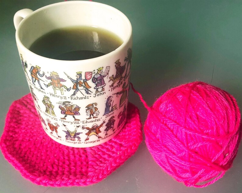 May include: A white mug with a historical figures design, filled with a green liquid, rests on a bright pink crocheted coaster. A ball of bright pink yarn sits nearby, suggesting a crafting theme.