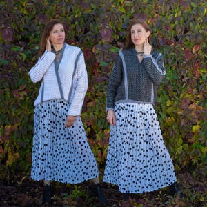 May include: Two women wearing matching outfits. Each outfit includes a white and gray sweater, a pleated skirt with black polka dots, and black boots. The sweaters have a V-neck design with gray trim. The background is a wall of green and red leaves.