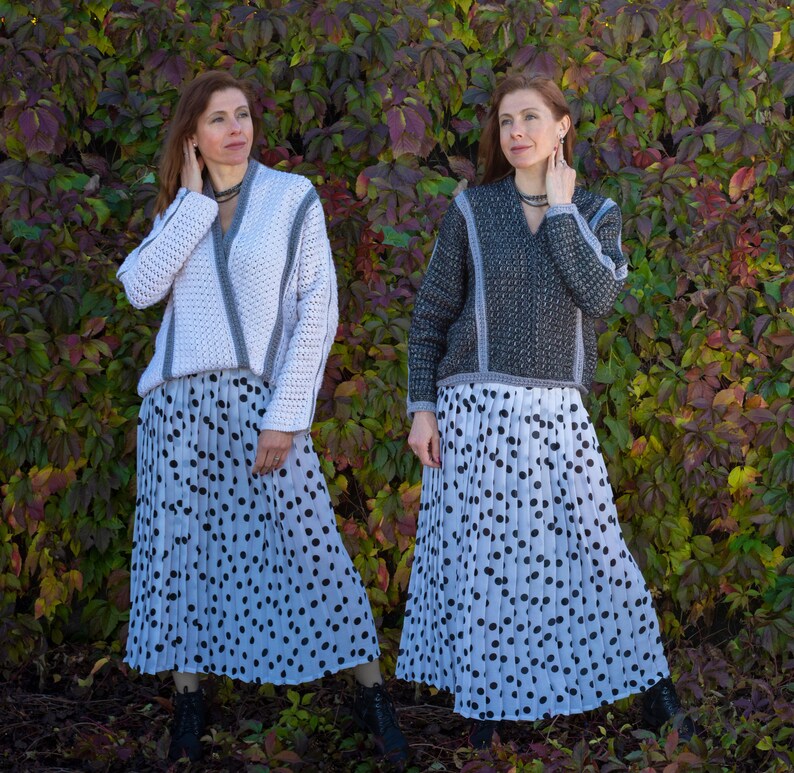 May include: Two women wearing matching outfits. Each outfit includes a white and gray sweater, a pleated skirt with black polka dots, and black boots. The sweaters have a V-neck design with gray trim. The background is a wall of green and red leaves.