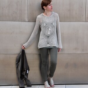 May include: A light beige, long-sleeved crochet hoodie with a circular pattern design and a tassel drawstring. The person is wearing olive green leggings and sandals, holding a black jacket.
