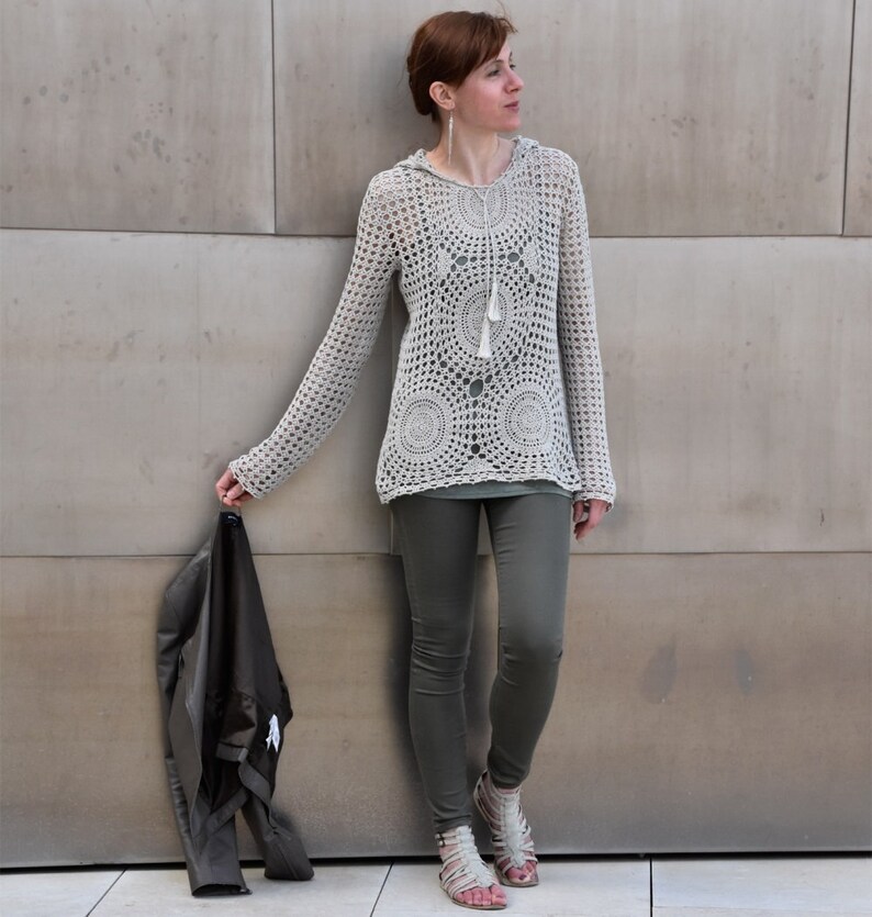 May include: A light beige, long-sleeved crochet hoodie with a circular pattern design and a tassel drawstring. The person is wearing olive green leggings and sandals, holding a black jacket.