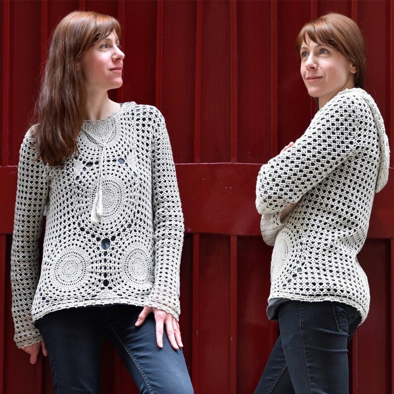 May include: A cream-colored, open-weave crochet sweater with a hood and long sleeves. The sweater features circular patterns on the front and a drawstring neckline. The model is wearing dark blue jeans. The background is a solid red.