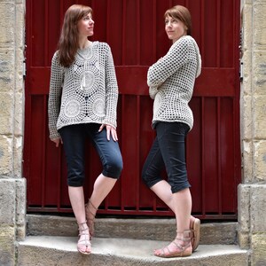 May include: Two women wearing light beige crochet sweaters with circular patterns and long sleeves, paired with dark capri pants and sandals. The background features a red door and stone wall.