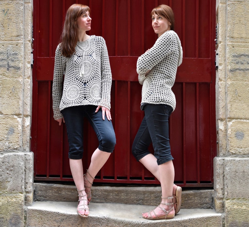 May include: Two women wearing light beige crochet sweaters with circular patterns and long sleeves, paired with dark capri pants and sandals. The background features a red door and stone wall.