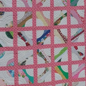 May include: A handmade quilt with a grid pattern of squares. Each square contains a white background with a curved design and a colorful arc of fabric. The quilt has a pink border and a white backing with a subtle pattern.