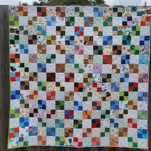 May include: A patchwork quilt with a grid pattern of squares in various colors and patterns. The squares include floral, geometric, and solid fabrics. The quilt is hanging on a wooden fence.