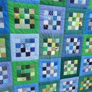 May include: A handmade quilt with a patchwork design. The quilt features squares of various blue and green fabrics, each framed by a contrasting border. The squares are arranged in a grid pattern, creating a visually appealing textile.