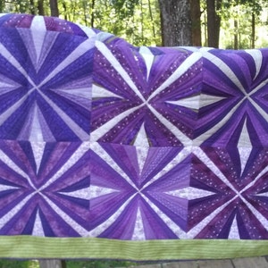 May include: A handmade quilt featuring a geometric starburst pattern in shades of purple and white. The quilt has a light green border and is displayed outdoors, showcasing its vibrant colors and intricate design. The quilt is a cozy home decor item.