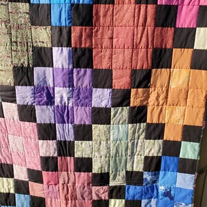 May include: A handmade quilt with a patchwork design. The quilt features a grid pattern of squares in various colors, including blue, pink, orange, purple, and black. The squares are made from different fabrics, creating a textured and visually interesting piece.
