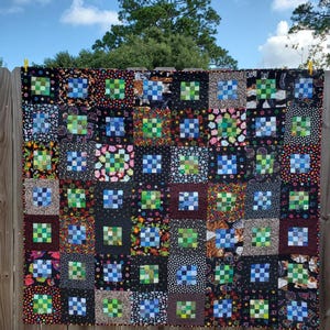 May include: A handmade quilt with a patchwork design, featuring squares of various patterns and colors. The quilt is primarily black with squares of blue, green, and other colorful fabrics. It is hanging on a wooden fence against a blue sky.