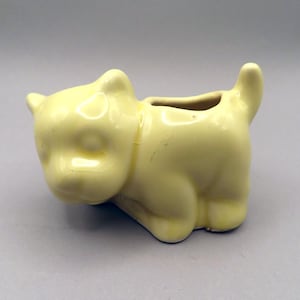 May include: A light yellow ceramic cat-shaped planter. The planter has a small opening on top for plants. The cat is in a crouching position with its tail up. The ceramic has a glossy finish.
