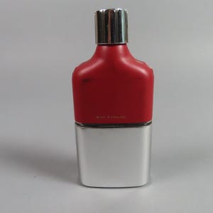 May include: A rectangular perfume bottle with a silver base and a red upper section. The bottle has a silver cap and the text "MADE IN ENGLAND" is visible. The bottle is on a gray background.