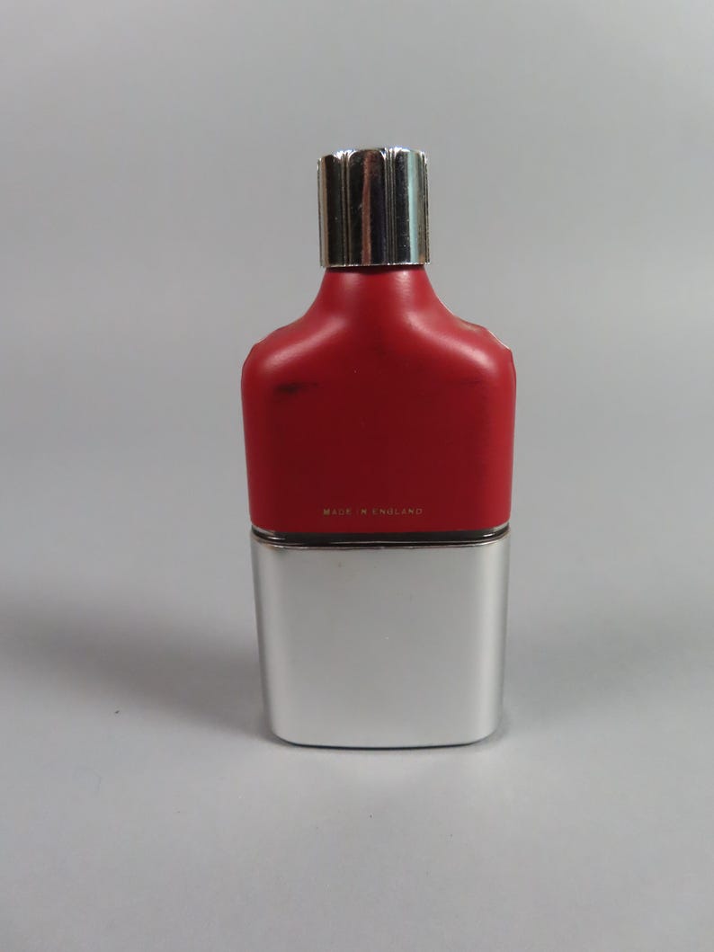 May include: A rectangular perfume bottle with a silver base and a red upper section. The bottle has a silver cap and the text "MADE IN ENGLAND" is visible. The bottle is on a gray background.