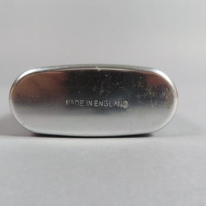 May include: A silver-colored, oval-shaped flask with rounded edges. The text "MADE IN ENGLAND" is imprinted on the flask's surface. The flask has a slightly worn appearance.