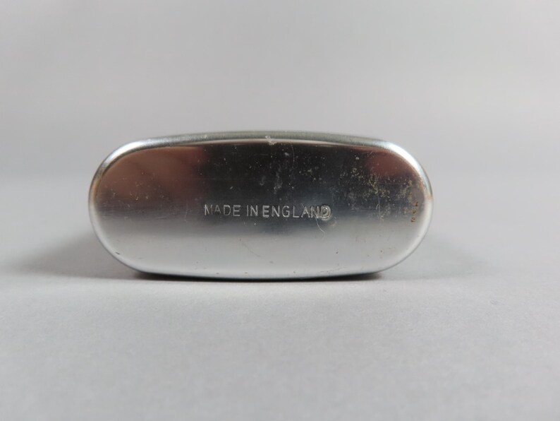 May include: A silver-colored, oval-shaped flask with rounded edges. The text "MADE IN ENGLAND" is imprinted on the flask's surface. The flask has a slightly worn appearance.