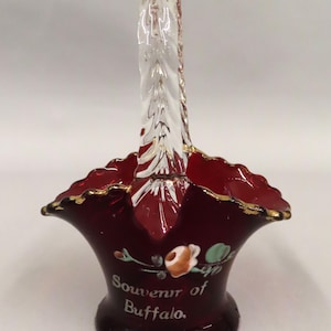 May include: A ruby red glass basket with a clear twisted handle. The rim is trimmed with gold. The words "Souvenir of Buffalo" are printed on the side, along with a floral design. A decorative piece.