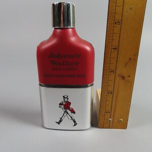 May include: A red and white Johnnie Walker Red Label bottle with a silver cap. The bottle has the brand name printed on it and a graphic of a man in a red coat. A wooden ruler is next to the bottle.