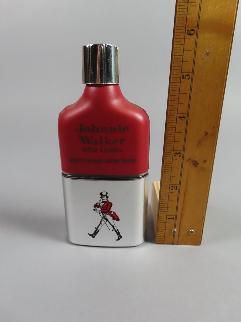 May include: A red and white Johnnie Walker Red Label bottle with a silver cap. The bottle has the brand name printed on it and a graphic of a man in a red coat. A wooden ruler is next to the bottle.