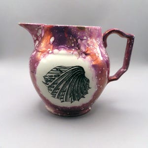 May include: A ceramic pitcher with a handle and spout. The pitcher has a white base with a black seashell design. The exterior is a mix of purple, copper, and white speckled glaze. The handle and spout are also glazed.