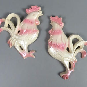 May include: Two wall-mounted rooster plaques featuring a cream-colored body with soft pink accents on the comb, wattle, and tail feathers. Each rooster is depicted in profile with a textured dotted pattern on the chest and curved tail feathers.