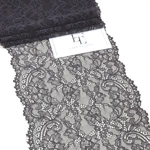May include: A roll of black lace fabric featuring a detailed floral and paisley pattern with scalloped edges. A white rectangular tag is tucked into the fabric, displaying the text LACE TO LOVE with a stylised logo above the words.
