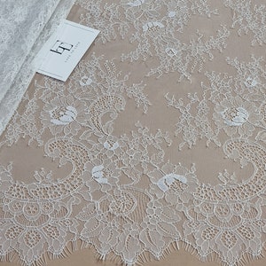 May include: White floral lace fabric with a scalloped edge. The lace features a delicate pattern of flowers and leaves. A folded section of the fabric is visible on the left, and a tag with the text "Lace to Love" is on the lower left.